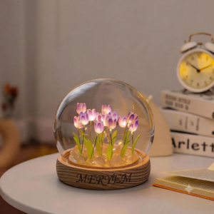 Une lampe de chevet en forme de dôme en verre contient un bouquet de tulipes violettes et blanches éclairées par des LED, posé sur une base en bois ronde gravée du prénom "MERYEM". La lampe est sur une table blanche à côté d'un réveil vintage et d'une pile de livres.